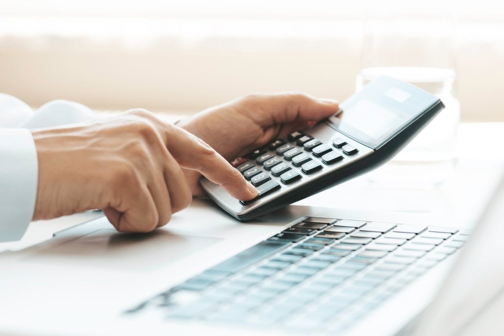 Man calculating customer acquisition cost on a calculator in front of a laptop - Shift4Shop